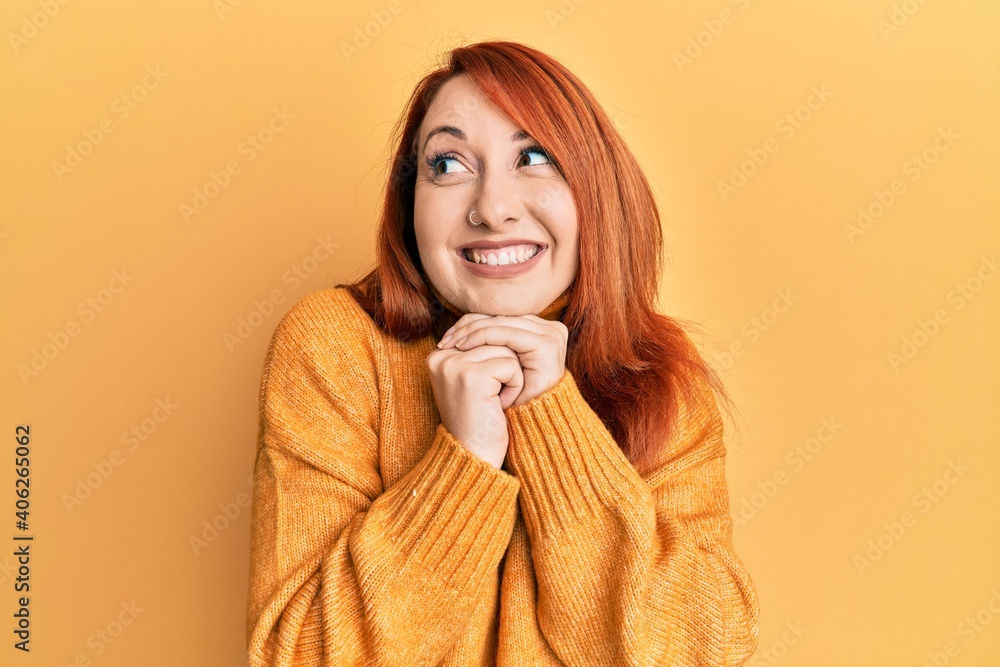 Beautiful redhead woman wearing casual winter sweater over yellow background laughing nervous and excited with hands on chin looking to the side Beautiful redhead woman wearing casual winter sweater over yellow background laughing nervous and excited with hands on chin looking to the side