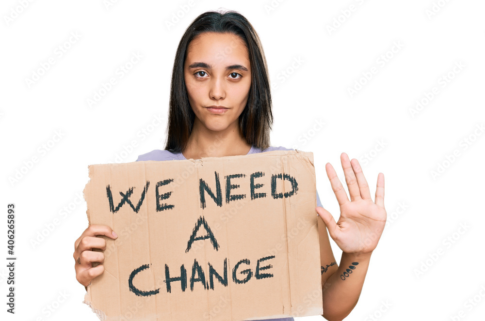 Young hispanic woman holding we need a change banner with open hand doing stop sign with serious and confident expression, defense gesture Young hispanic woman holding we need a change banner with open hand doing stop sign with serious and confident expression, defense gesture