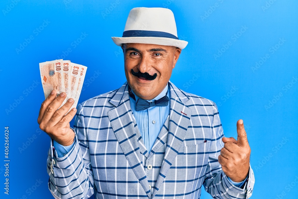 Mature middle east man with mustache wearing elegant vintage style holding pounds banknotes smiling happy pointing with hand and finger to the side Mature middle east man with mustache wearing elegant vintage style holding pounds banknotes smiling happy pointing with hand and finger to the side