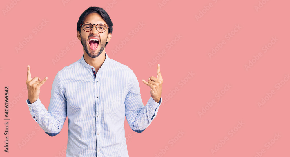Handsome hispanic man wearing business shirt and glasses shouting with crazy expression doing rock symbol with hands up. music star. heavy music concept.