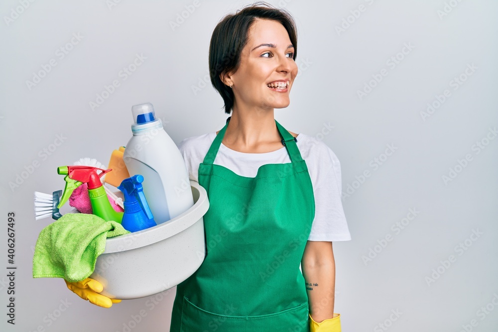 Young brunette woman with short hair wearing apron holding cleaning products looking away to side with smile on face, natural expression. laughing confident. Young brunette woman with short hair wearing apron holding cleaning products looking away to side with smile on face, natural expression. laughing confident.