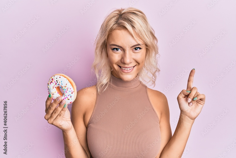 Young blonde girl holding donut smiling with an idea or question pointing finger with happy face, number one Young blonde girl holding donut smiling with an idea or question pointing finger with happy face, number one