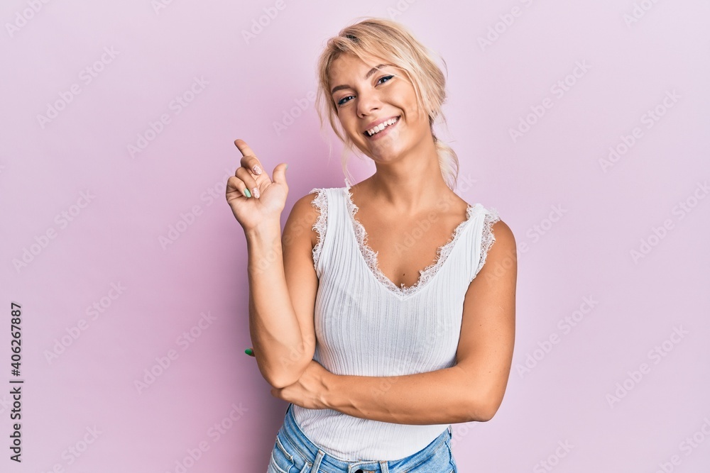 Young blonde girl wearing casual clothes with a big smile on face, pointing with hand and finger to the side looking at the camera.