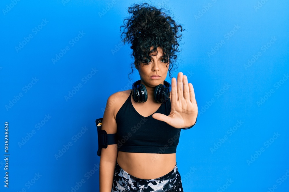 Young hispanic woman wearing gym clothes and using headphones doing stop sing with palm of the hand. warning expression with negative and serious gesture on the face.