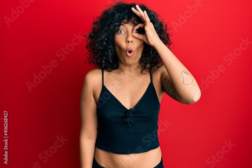 Young hispanic woman wearing casual style with sleeveless shirt doing ok gesture shocked with surprised face, eye looking through fingers. unbelieving expression.