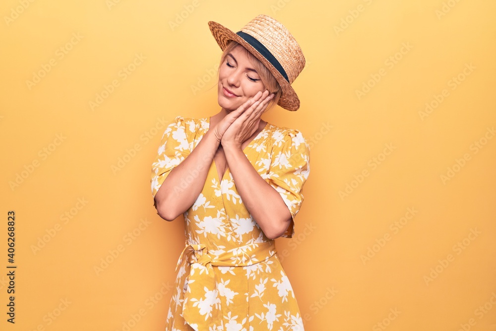 Beautiful blonde woman on vacation wearing summer hat and dress over yellow background sleeping tired dreaming and posing with hands together while smiling with closed eyes. Beautiful blonde woman on vacation wearing summer hat and dress over yellow background sleeping tired dreaming and posing with hands together while smiling with closed eyes.