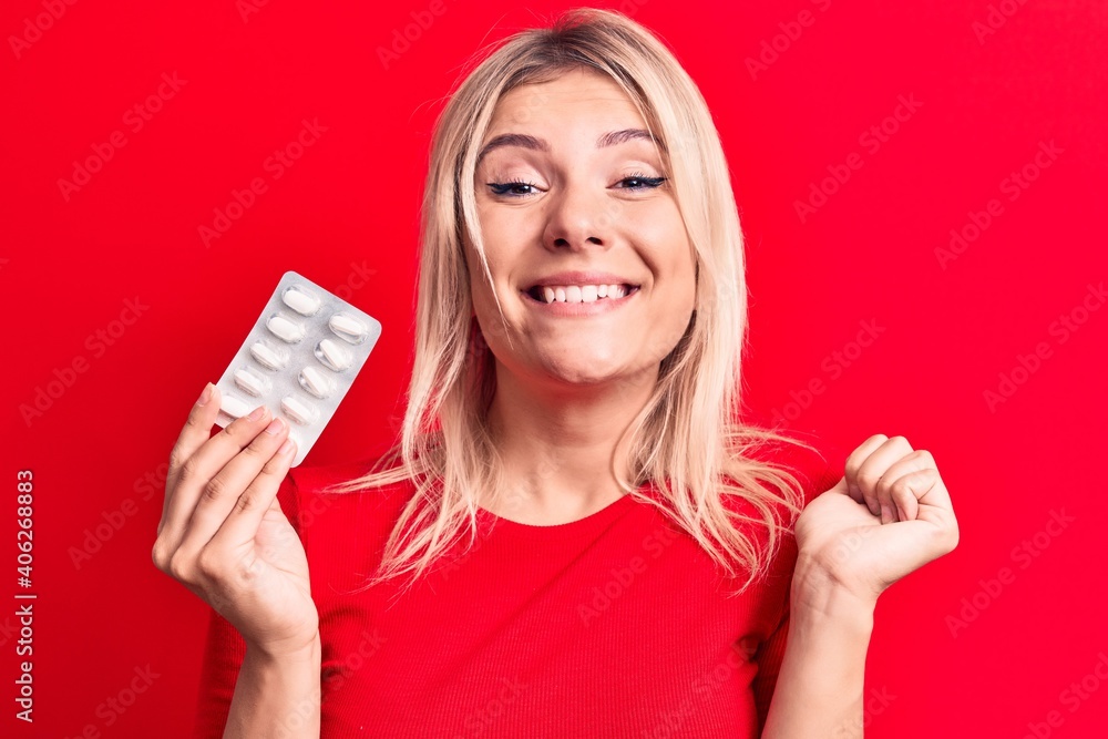 Young beautiful blonde woman holding pills standing over isolated red background screaming proud, celebrating victory and success very excited with raised arm Young beautiful blonde woman holding pills standing over isolated red background screaming proud, celebrating victory and success very excited with raised arm
