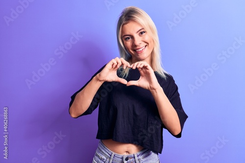 Young beautiful blonde woman wearing casual black t-shirt standing over purple background smiling in love doing heart symbol shape with hands. Romantic concept.