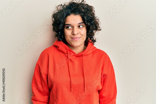 Young hispanic woman with curly hair wearing casual sweatshirt smiling looking to the side and staring away thinking.