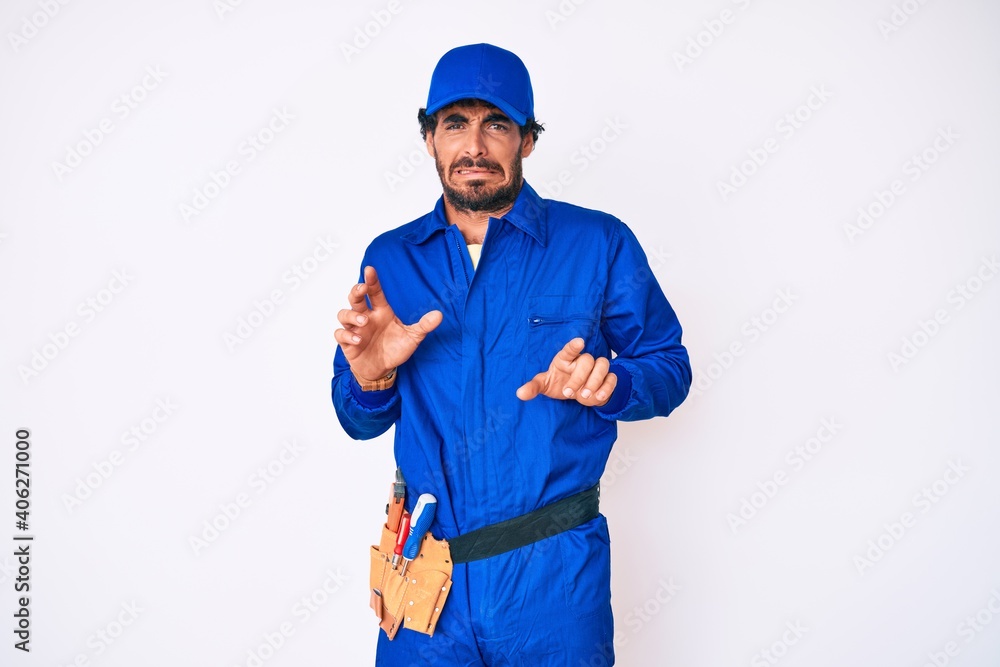 Handsome young man with curly hair and bear weaing handyman uniform disgusted expression, displeased and fearful doing disgust face because aversion reaction.