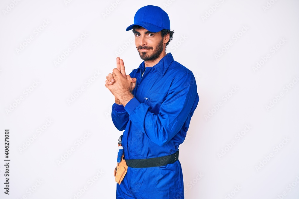 Handsome young man with curly hair and bear weaing handyman uniform holding symbolic gun with hand gesture, playing killing shooting weapons, angry face
