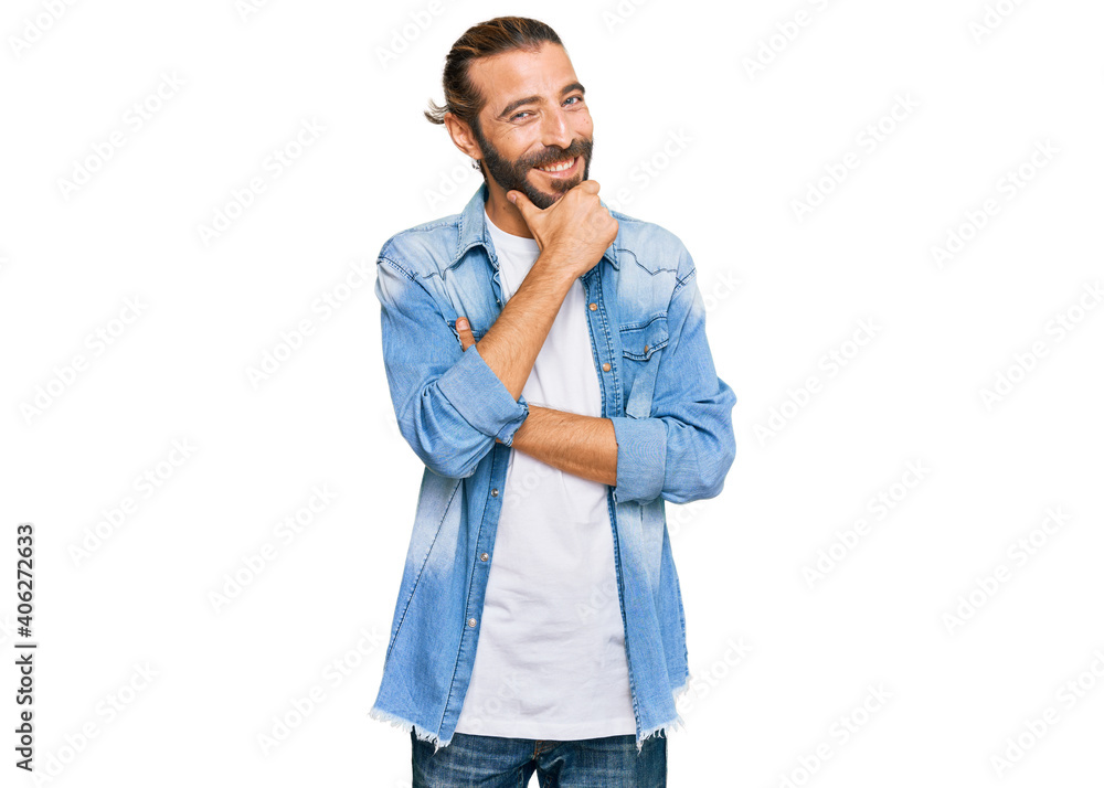 Attractive man with long hair and beard wearing casual denim jacket looking confident at the camera smiling with crossed arms and hand raised on chin. thinking positive. Attractive man with long hair and beard wearing casual denim jacket looking confident at the camera smiling with crossed arms and hand raised on chin. thinking positive.