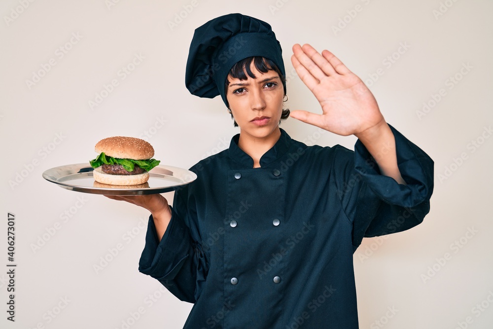 Beautiful brunettte woman chef holding burguer with open hand doing stop sign with serious and confident expression, defense gesture Beautiful brunettte woman chef holding burguer with open hand doing stop sign with serious and confident expression, defense gesture