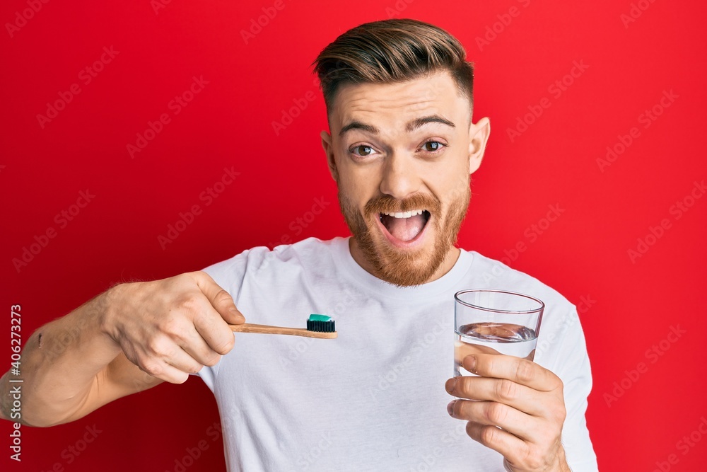 Young redhead man holding toothbrush with toothpaste and glass of water celebrating crazy and amazed for success with open eyes screaming excited. Young redhead man holding toothbrush with toothpaste and glass of water celebrating crazy and amazed for success with open eyes screaming excited.