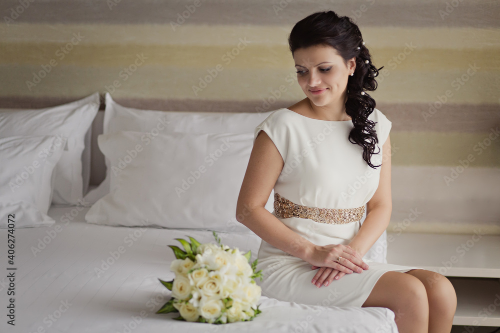 Pretty young Bride in simple wedding dress with her wedding bouquet. Black-haired woman with wedding hair-style with a long tail. Boudoir morning of the bride in modern room Pretty young Bride in simple wedding dress with her wedding bouquet. Black-haired woman with wedding hair-style with a long tail. Boudoir morning of the bride in modern room