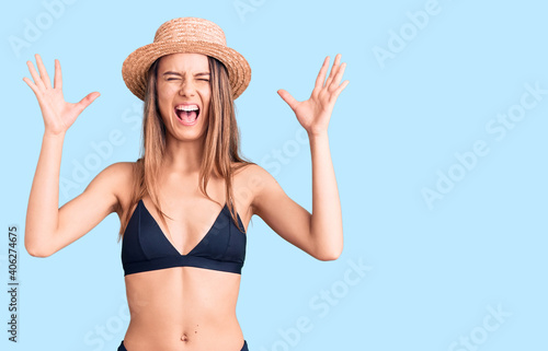 Young beautiful girl wearing bikini and hat celebrating mad and crazy for success with arms raised and closed eyes screaming excited. winner concept
