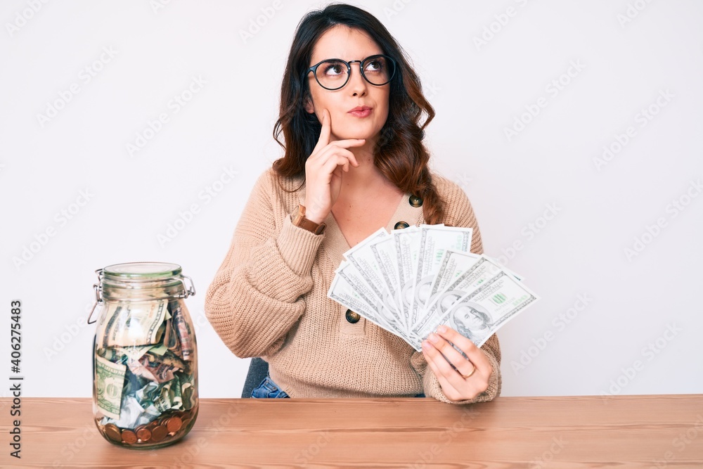 Young beautiful brunette woman holding jar with savings holding dollars serious face thinking about question with hand on chin, thoughtful about confusing idea Young beautiful brunette woman holding jar with savings holding dollars serious face thinking about question with hand on chin, thoughtful about confusing idea