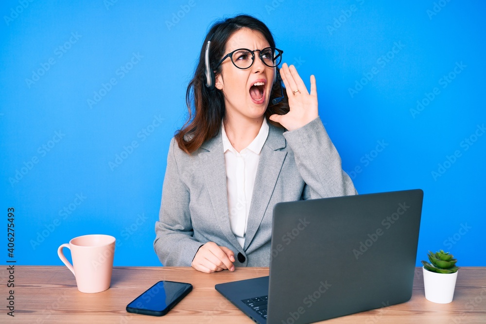 Young beautiful brunette woman working at the office wearing operator headset shouting and screaming loud to side with hand on mouth. communication concept.