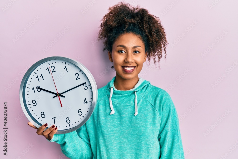 Beautiful african american woman with afro hair holding big clock looking positive and happy standing and smiling with a confident smile showing teeth Beautiful african american woman with afro hair holding big clock looking positive and happy standing and smiling with a confident smile showing teeth