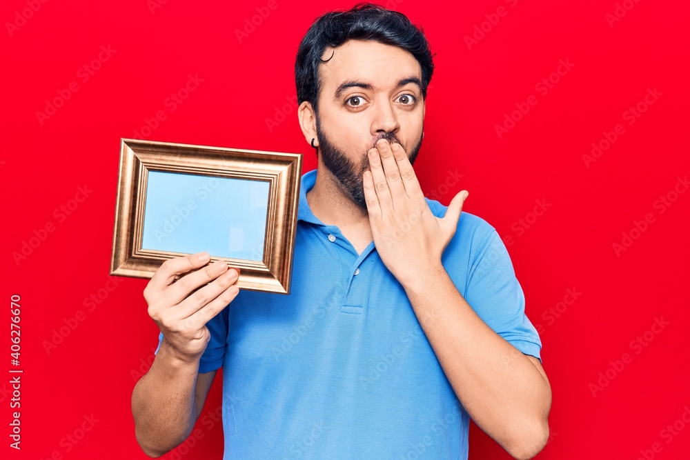 Young hispanic man holding empty frame covering mouth with hand, shocked and afraid for mistake. surprised expression Young hispanic man holding empty frame covering mouth with hand, shocked and afraid for mistake. surprised expression