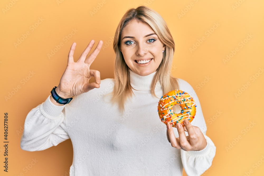 Young caucasian woman holding donut doing ok sign with fingers, smiling friendly gesturing excellent symbol Young caucasian woman holding donut doing ok sign with fingers, smiling friendly gesturing excellent symbol