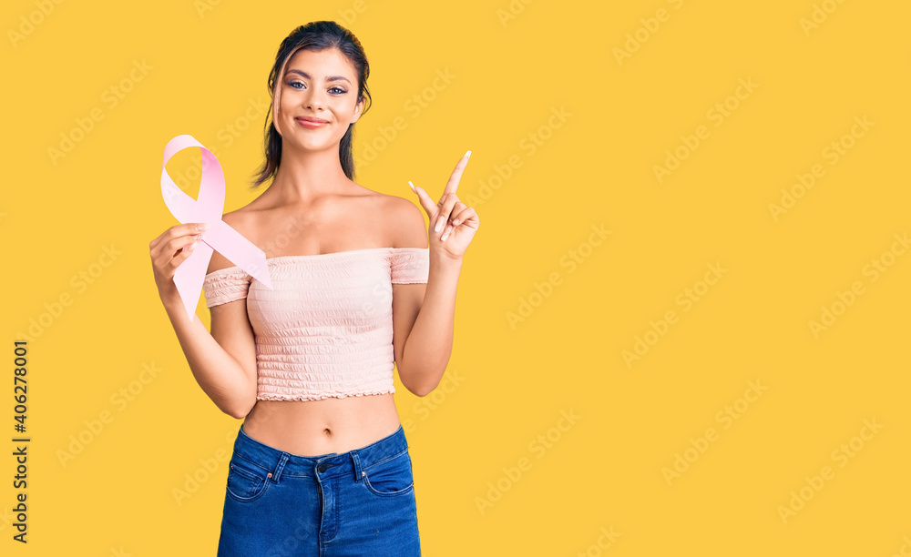 Young beautiful woman holding cancer awareness pink ribbon surprised with an idea or question pointing finger with happy face, number one Young beautiful woman holding cancer awareness pink ribbon surprised with an idea or question pointing finger with happy face, number one