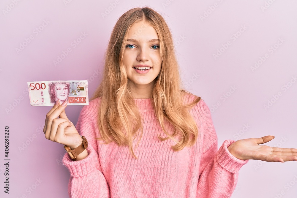 Beautiful young caucasian girl holding 500 swedish krona banknote celebrating achievement with happy smile and winner expression with raised hand Beautiful young caucasian girl holding 500 swedish krona banknote celebrating achievement with happy smile and winner expression with raised hand