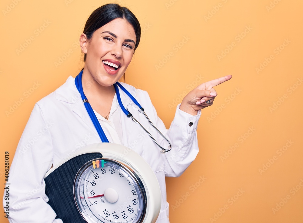 Young beautiful doctor woman wearing stethoscope and coat holding weighting machine smiling happy pointing with hand and finger to the side Young beautiful doctor woman wearing stethoscope and coat holding weighting machine smiling happy pointing with hand and finger to the side
