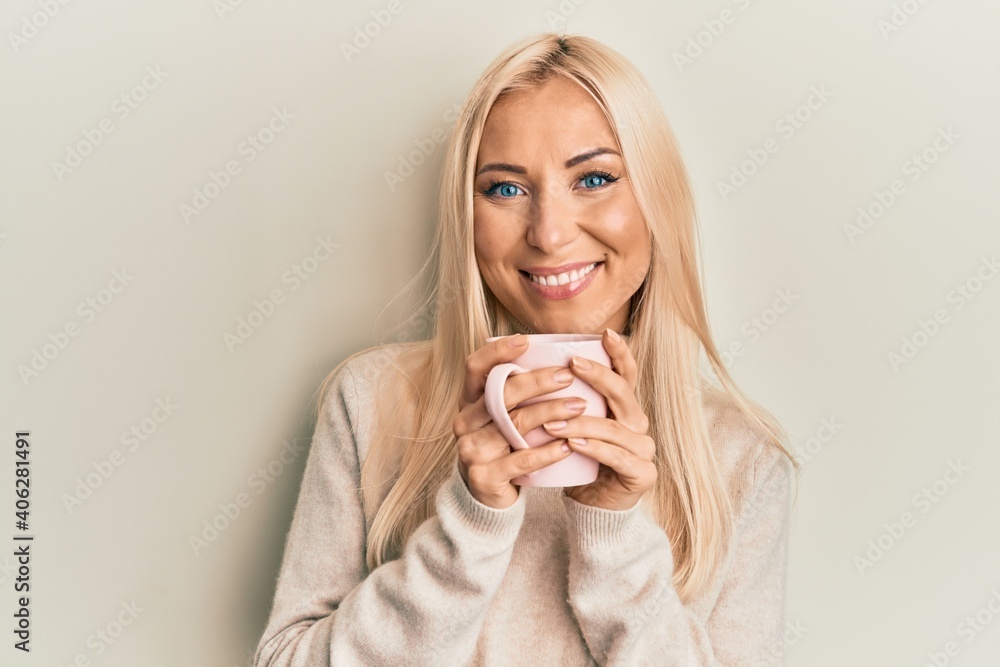 Young blonde woman drinking a cup of coffee smiling with a happy and cool smile on face. showing teeth. Young blonde woman drinking a cup of coffee smiling with a happy and cool smile on face. showing teeth.