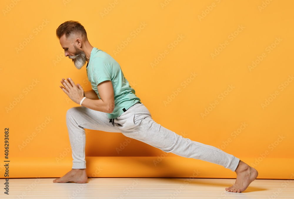 Portrait of an elderly yogi man in comfortable clothes exercising against a yellow wall doing yoga or pilates. Standing in the Virabhadrasana warrior pose. stretching Portrait of an elderly yogi man in comfortable clothes exercising against a yellow wall doing yoga or pilates. Standing in the Virabhadrasana warrior pose. stretching
