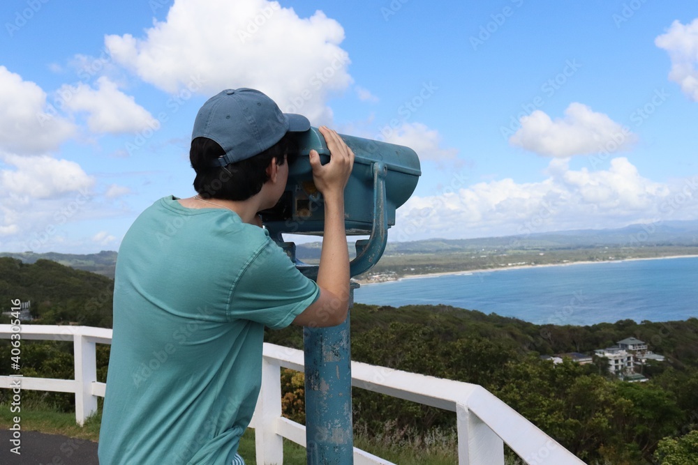 Obraz premium A young male tourist wearing a cap and a green t-shirt observing the stunning view of Cape Byron bay in Australia from a lookout binoculars.