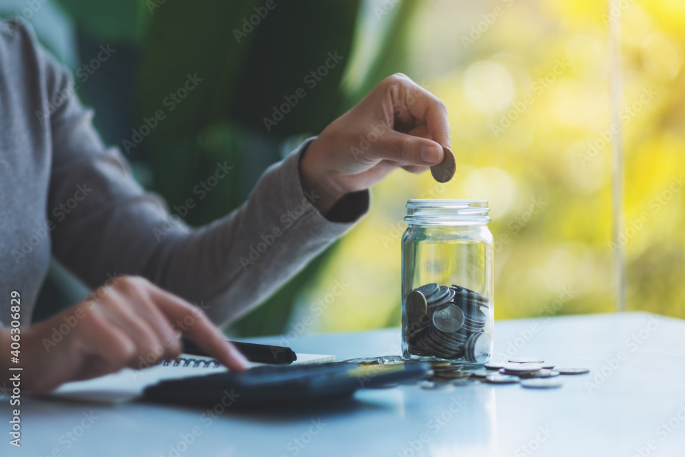 Closeup image of a woman putting coins in a glass jar and calculating on calculator for saving money and financial concept