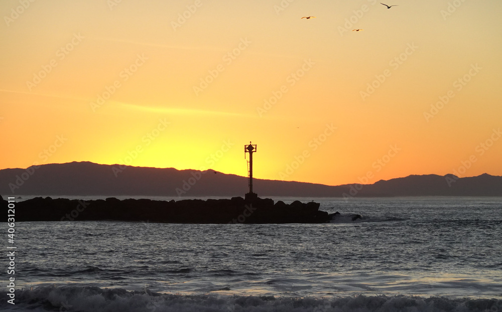 Sunset over ocean, rocky break waters, beacon and birds along coast of California