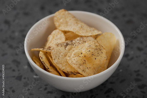 White bowl with organic potato chips sprinkled with fresh ground black pepper on concrete background