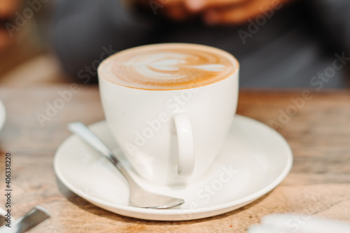 Closeup shot of white cup filled with aromatic coffee with heart shape latte art foam on wood table near window with light shade on tabletop at cafe with spoon besides it