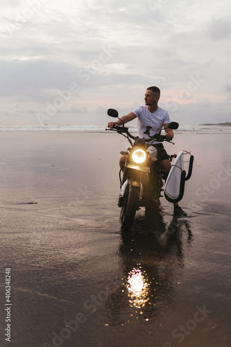 Surfer rides on motorbike with surfboard at sunset ocean beach. Bali island, Indonesia