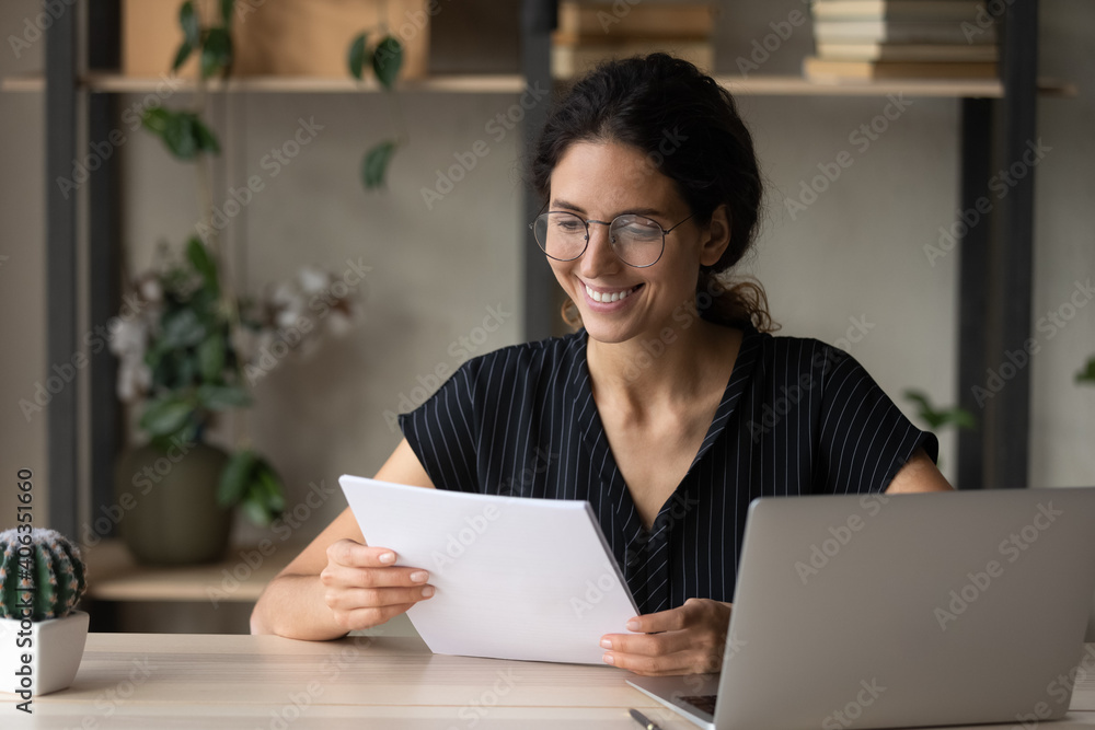 Smiling millennial female in spectacles read paper documents work on computer online at home office. Happy young Caucasian woman in glasses consider paperwork use laptop for distant job or studying. Smiling millennial female in spectacles read paper documents work on computer online at home office. Happy young Caucasian woman in glasses consider paperwork use laptop for distant job or studying.
