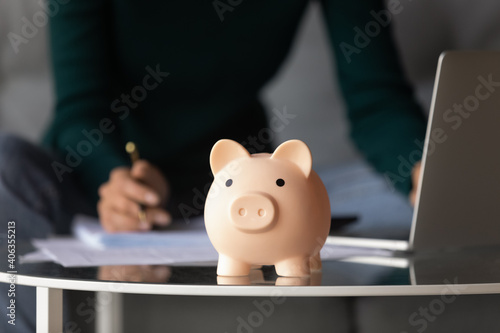 Crop close up of piggy bank on table, woman manage family budget pay online on laptop on background. Female take care of expenses expenditures, make investment or money saving. Planning concept.
