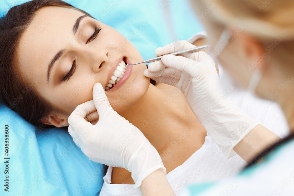 Young female patient visiting dentist office.Beautiful woman with healthy straight white teeth sitting at dental chair with open mouth during oral checkup while doctor working at teeth Young female patient visiting dentist office.Beautiful woman with healthy straight white teeth sitting at dental chair with open mouth during oral checkup while doctor working at teeth