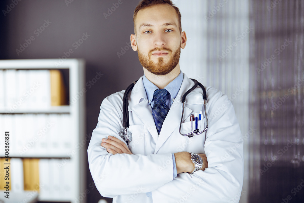 Red-bearded doctor standing straight in clinic near his working place. Portrait of physician. Medicine and healthcare concept Red-bearded doctor standing straight in clinic near his working place. Portrait of physician. Medicine and healthcare concept