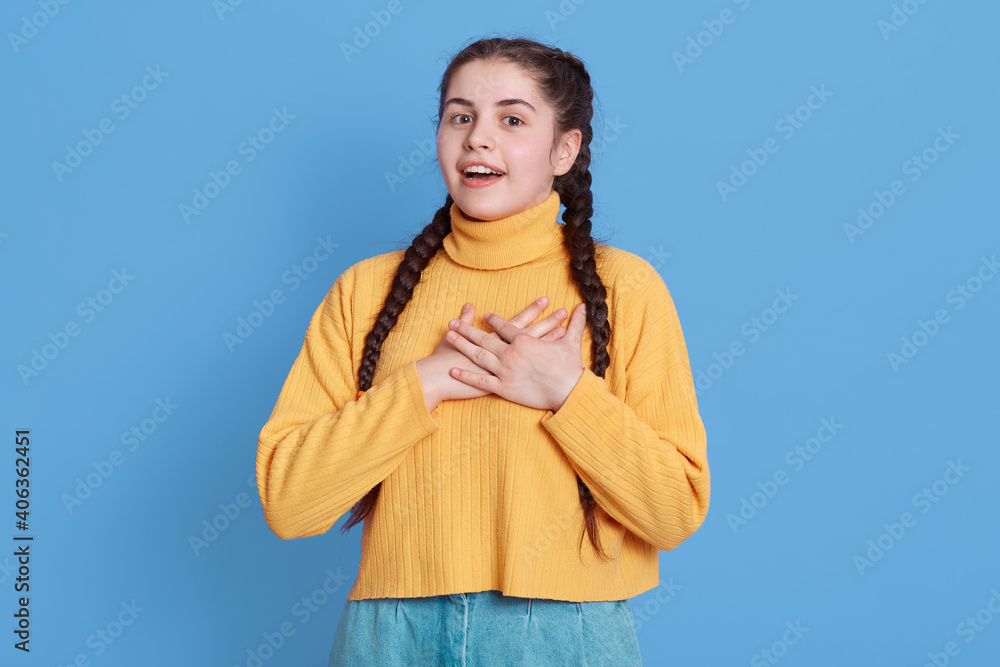 Touched thankful European woman keeps hands on heart, expresses gratitude and kindness, smiles happily, wears casual yellow sweater and jeans, thanks for something, poses against blue background. Touched thankful European woman keeps hands on heart, expresses gratitude and kindness, smiles happily, wears casual yellow sweater and jeans, thanks for something, poses against blue background.