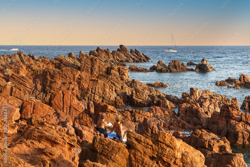 Sunset over Esterel mediterranean red rocks coast, French Riviera, Cote d Azur, Provence, France. Boats in the sea, back view of a couple relaxing on the rocks. Holidays in France.