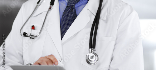Unknown male doctor iusing tablet computer in his hands, while standing in clinic. Medicine concept during Coronavirus pandemic
