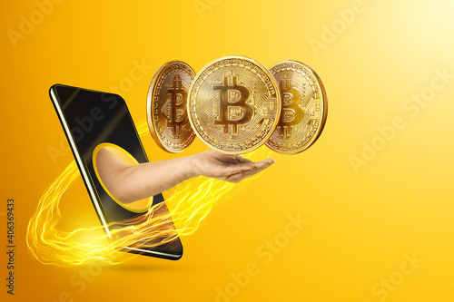 The hand crawling out through the smartphone holds, offers bitcoin coins, yellow background. Crypto currency concept, blockchain technology, money of the future. Copy space. photo