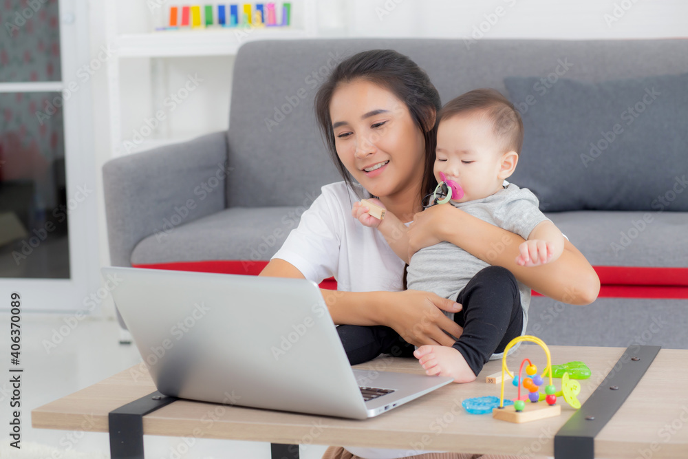 Young asian mother working on laptop computer while care daughter girl at home, mom and baby girl sitting using notebook video call, parent and bonding, indoors, family and business concept. Young asian mother working on laptop computer while care daughter girl at home, mom and baby girl sitting using notebook video call, parent and bonding, indoors, family and business concept.