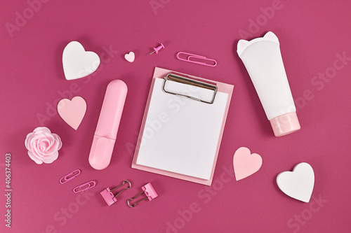 Clipboard with empty paper for text surrounded by hearts, beauty producs and stationery items on dark pink background