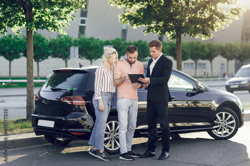 A young salesman shows a new car to customers. Happy couple, man and woman buy a new car. Young people sign documents to buy a car