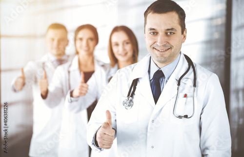 Group of professional doctors are standing as a team with thumbs up in a sunny hospital office, ready to help their patients. Modern medical help, insurance in health care and medicine concept