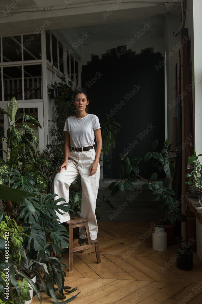 Young woman gardener with natural makeup stands in a greenhouse, surrounded by plants and pots. Woman is wearing white trousers. Love plants, hobby, home gardening concept, freelance, small business. Young woman gardener with natural makeup stands in a greenhouse, surrounded by plants and pots. Woman is wearing white trousers. Love plants, hobby, home gardening concept, freelance, small business.