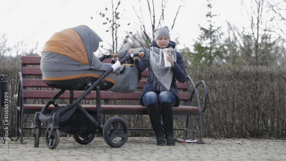 A young mother is walking in a Park bench, rocking a stroller and drinking tea from a thermos. Family, parenthood concept - happy mother with stroller in park. Woman with baby outdoor. A young mother is walking in a Park bench, rocking a stroller and drinking tea from a thermos. Family, parenthood concept - happy mother with stroller in park. Woman with baby outdoor.